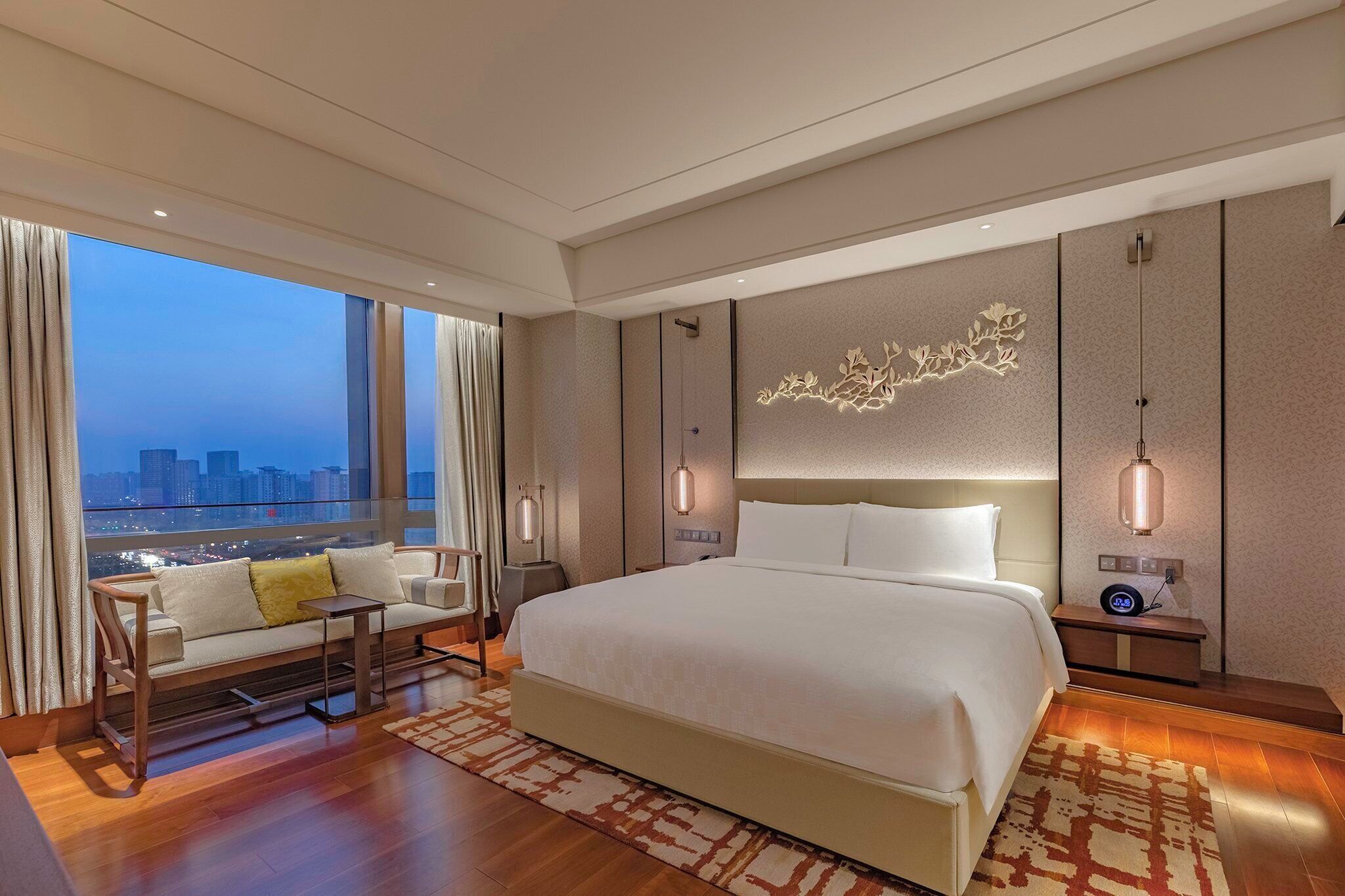A modern hotel room with a large bed, a seating area with a sofa and armchairs, a city view through large windows, and decorative wall panels.