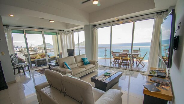 A spacious living area with a large sofa, a coffee table, and a TV. There is a balcony with a view of the sea and a dining area with chairs. - Mazatlán Solaria Oceanview Condo with Pool 704 (Mazatlan)