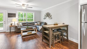Uma sala de estar moderna com sofá, mesa de centro e mesa de jantar. Há um ventilador de teto, uma janela com vista para o verde e uma geladeira.