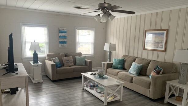A living room with two beige sofas, a white coffee table, a ceiling fan, and a framed picture on the wall. - Beach House with Pool - Pet Friendly (North Myrtle Beach)