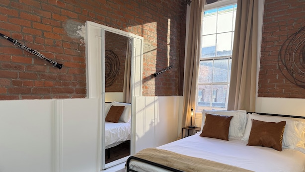 A bedroom with a brick wall, a bed with white and brown bedding, a window with curtains, and a small bedside table with a lamp.