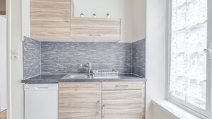 A modern kitchen with wooden cabinets, a grey tiled backsplash, a dishwasher, and a window with a white curtain. - Ermine - Intramuros proche plage - St Malo (Saint-Malo)