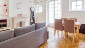 A living room with a grey sofa, a dining table with chairs, a radiator, a clock, and a window. - Ermine - Intramuros proche plage - St Malo (Saint-Malo)