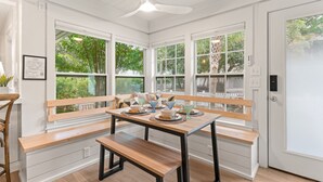 Una cocina con mesa y banco de madera, ventanas amplias con vistas a follaje, y armarios blancos.