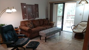 A living room with a brown sofa, a black leather chair, a patterned ottoman, and a wicker chair. There is a sliding glass door leading to an outdoor area. - Beautifully furnished condo at the Villages in McCormick Ranch Scottsdale, AZ (Scottsdale)