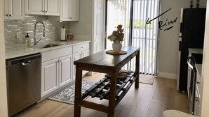 A kitchen with white cabinets, a stainless steel dishwasher, a wooden table with a vase of flowers, and a chandelier. - Home Base for New Smyrna Adventures (New Smyrna Beach)