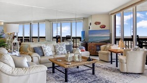 Un salón amplio con un sofá grande, una mesa de centro y un televisor. Hay ventanas grandes con vista al océano.
