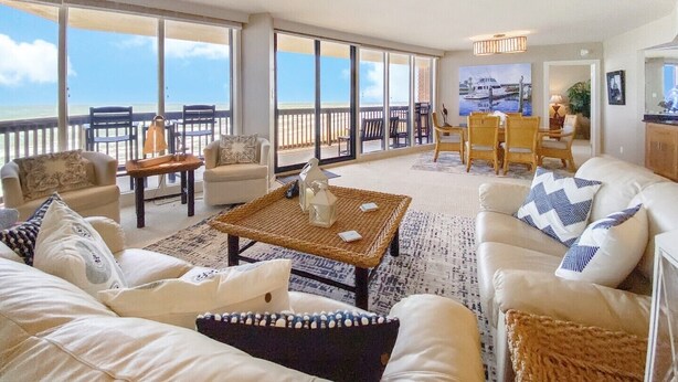 Amplia sala de estar con balcón vistas al mar, amueblada con sofás blancos, una mesa de centro y sillas de mimbre.
