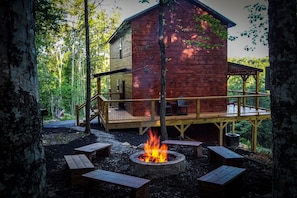 Relax on the wooden deck, surrounded by a forest, with a fire pit and benches for a tranquil retreat.