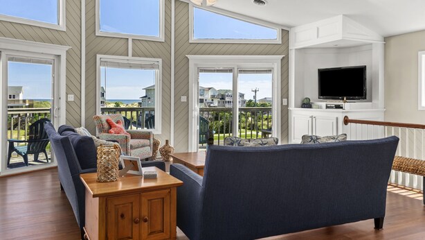 A spacious living room with a large window, a ceiling fan, and a flat-screen TV mounted on a white cabinet. - Beach Dugout: Semi-Oceanfront, Shared Pool (Nags Head)