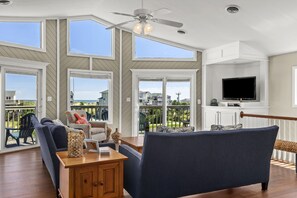 A spacious living room with a large window, a ceiling fan, and a flat-screen TV mounted on a white cabinet. - Beach Dugout: Semi-Oceanfront, Shared Pool (Nags Head)