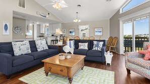 A spacious living room with a large ceiling fan, a wooden coffee table, and a view of the outdoors through large windows. - Beach Dugout: Semi-Oceanfront, Shared Pool (Nags Head)