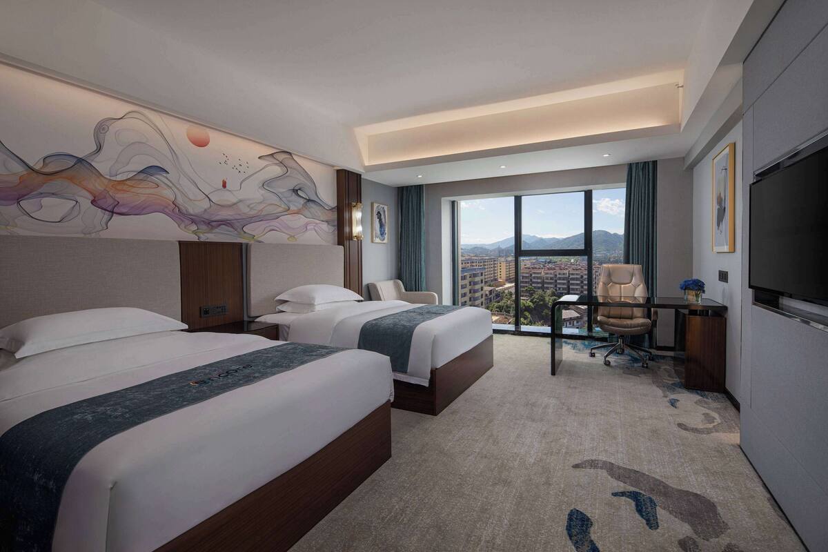Een hotelkamer met twee bedden, een bureau, een stoel, een televisie en een groot raam met uitzicht op de stad.