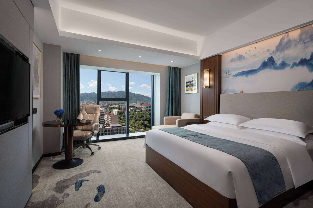 Een hotelkamer met een groot bed, een bureau met een stoel, een televisie en uitzicht op de stad en de bergen.