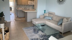 A living room with a glass coffee table, beige sofa, and a kitchen in the background. - Oceanfront condo with stunning views. Oahu’s last best kept secret (Waianae)