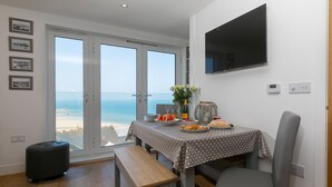 A dining area with a table set for two, a bench, and a view of the beach through large glass doors.