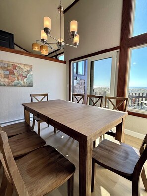 Un área de comedor con una mesa y sillas de madera, una araña, un ventanal con vistas y un mapa en la pared.