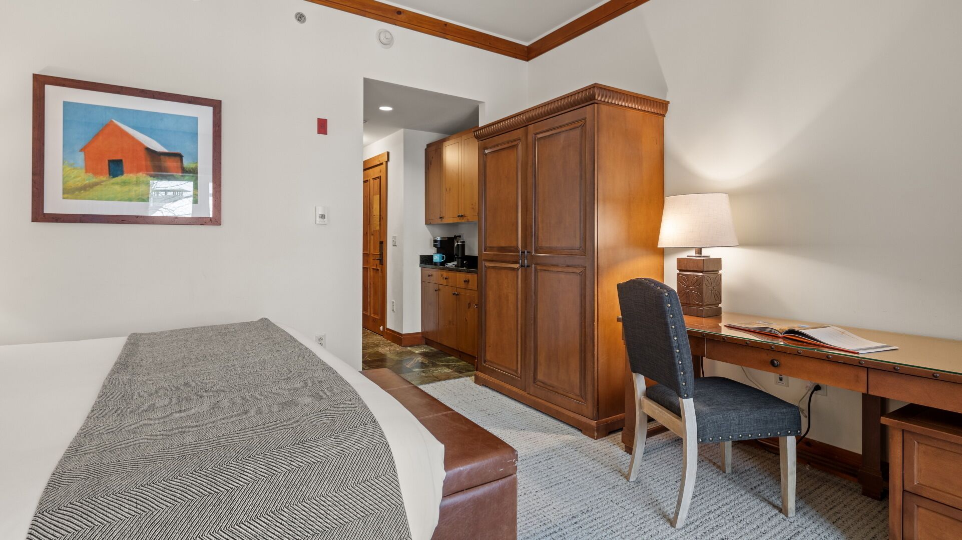 A hotel room with a bed, a desk with a chair, a lamp, and a painting of a barn.