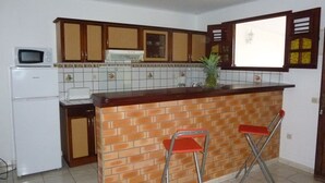 A kitchen with a tiled backsplash, wooden cabinets, a microwave, a refrigerator, and two red bar stools. - Apartment COLIBRI (Saint-Esprit)