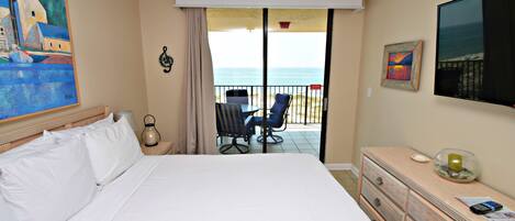 A hotel room with a large bed, a TV, a dresser, a chair, and a balcony with a view of the sea.