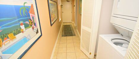A narrow hallway with a tiled floor leading to a door. There are framed pictures on the walls, a washing machine, and a door with a towel hanging on it.