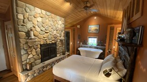 A bedroom with a stone fireplace, a large bed, a bathtub, and a ceiling fan. - Wild Goose Inn (East Lansing)