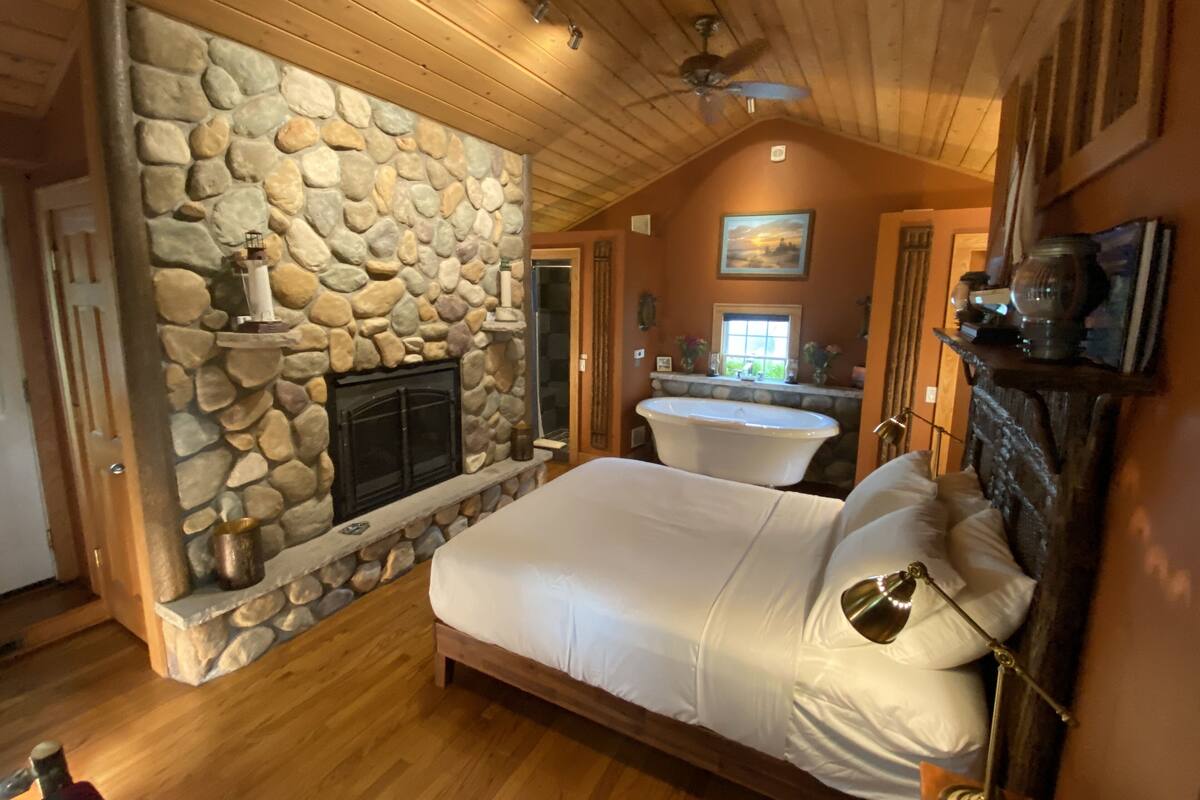 A bedroom with a stone fireplace, a large bed, a bathtub, and a ceiling fan.