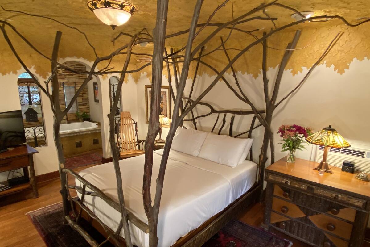 A rustic-style bedroom with a four-poster bed, a wooden nightstand, a lamp, and a chair.