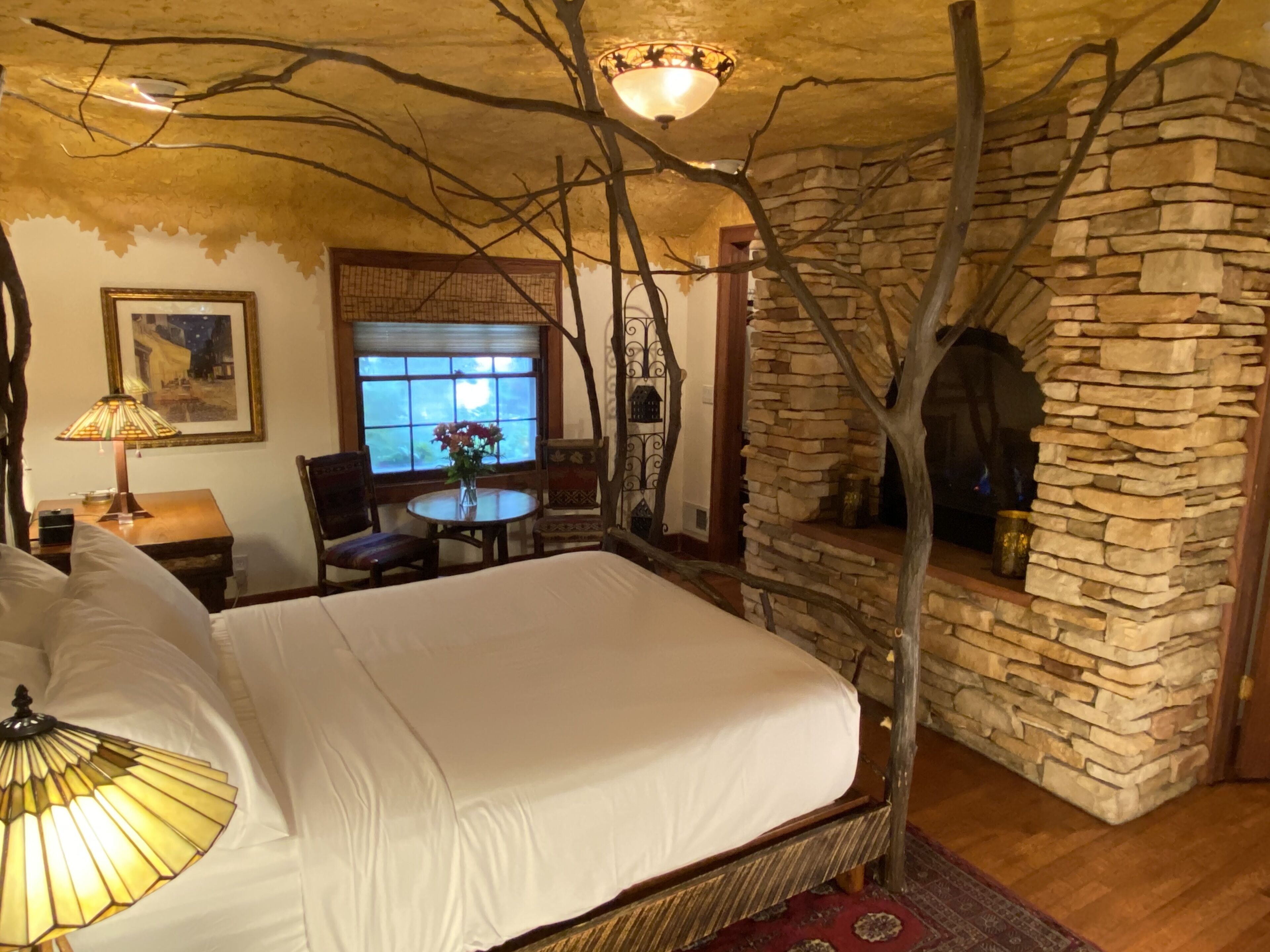Un dormitorio con chimenea de piedra, una cama con ropa de cama blanca, una mesita de noche de madera, una mesa con lámpara y una silla.