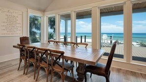 A dining area with a wooden table and chairs, large windows with a view of the beach and ocean, and a wall with a message.