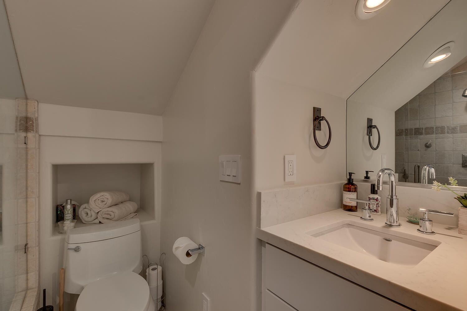 Une salle de bain moderne avec des toilettes, un lavabo et un miroir. On trouve un porte-serviettes au-dessus des toilettes et une étagère avec des serviettes.