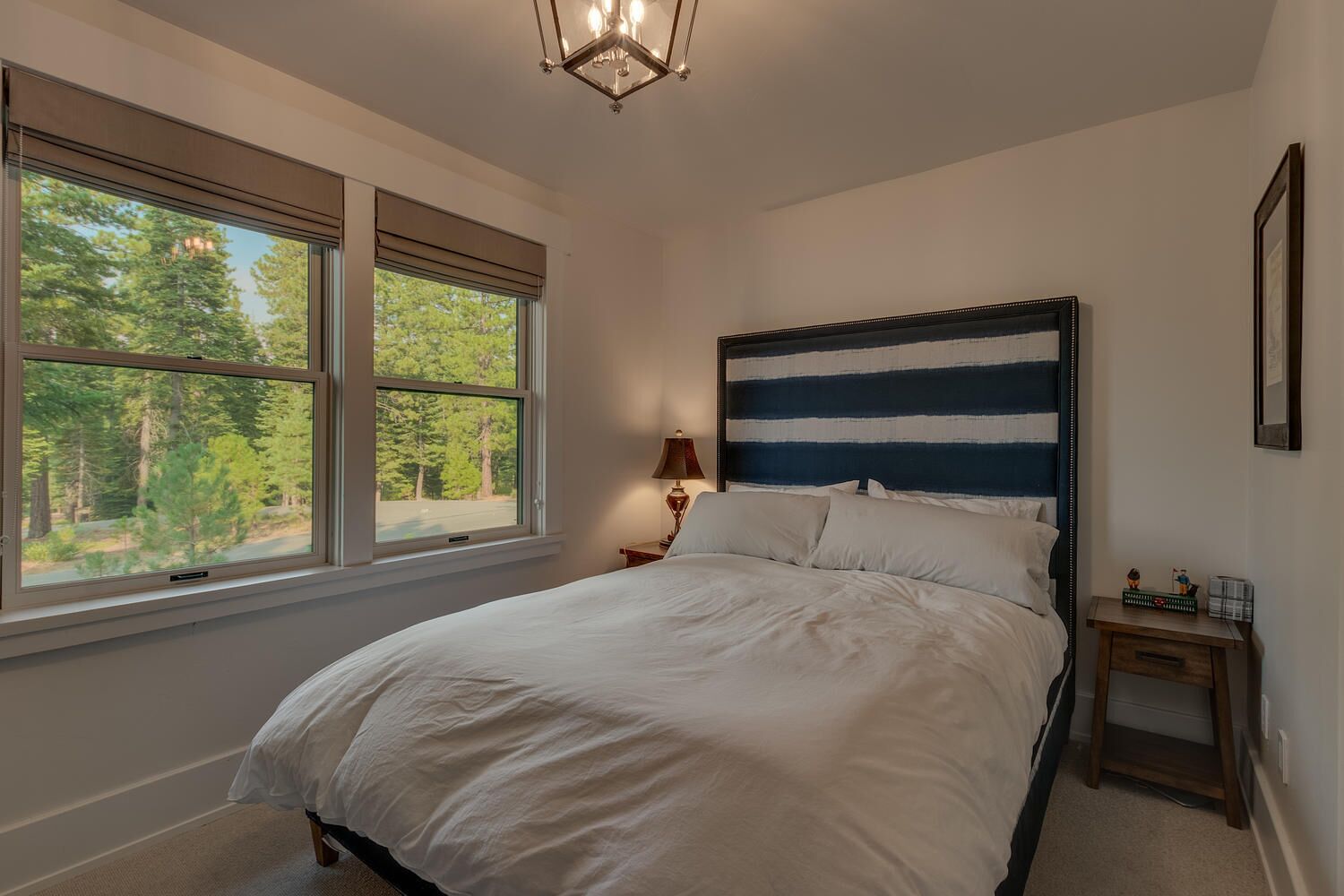 A bedroom with a large bed, two windows with blinds, a nightstand, a lamp, and a view of trees outside.
