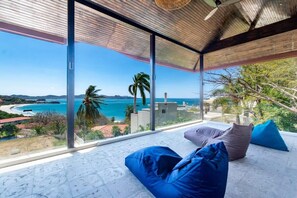 Une chambre moderne avec de grandes fenêtres en verre donnant sur la côte, dotée d’un plafond en bois et agrémentée de quelques coussins.