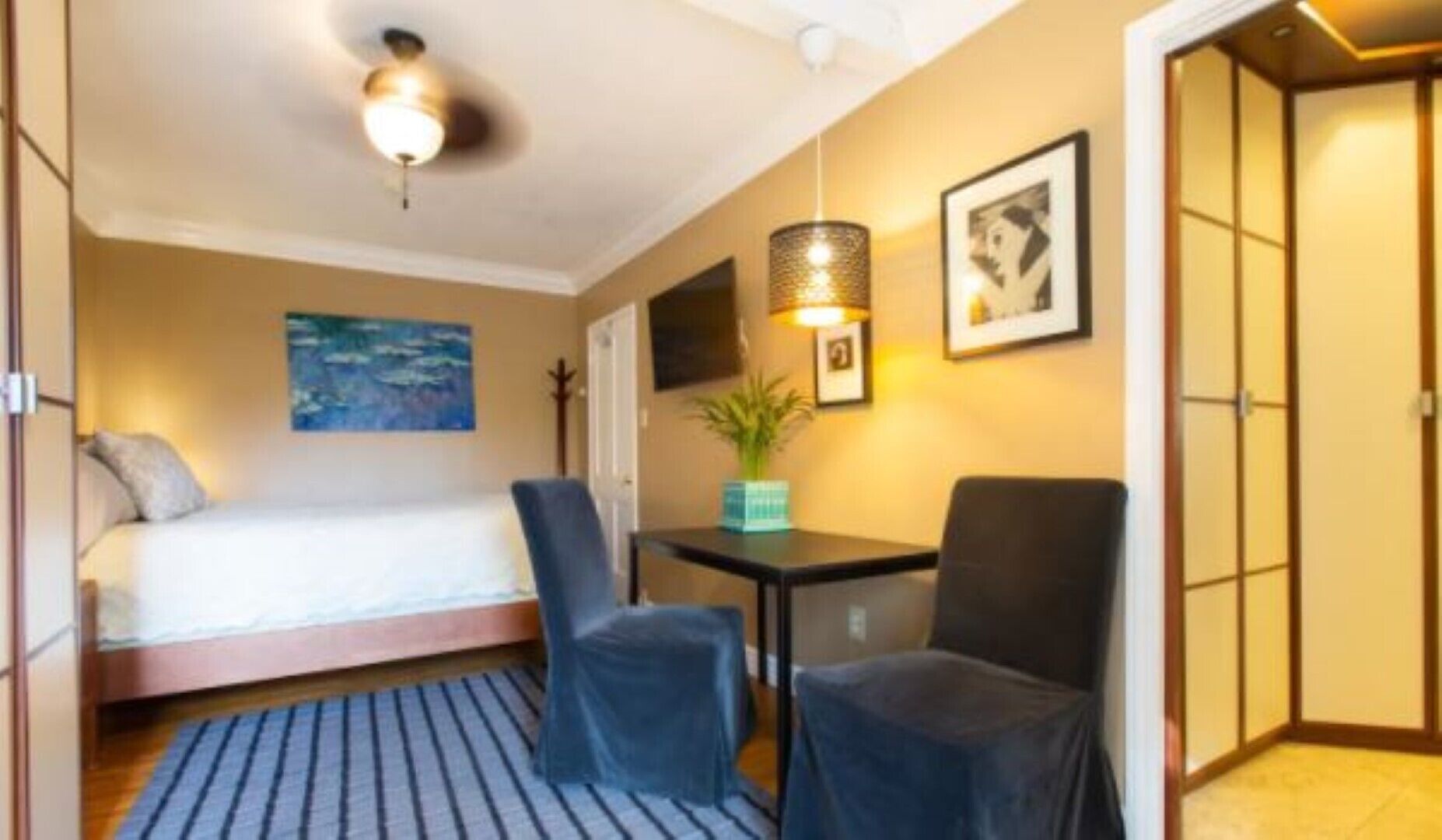 A hotel room with a bed, a desk with two chairs, a ceiling fan, a painting, and a wall-mounted TV.