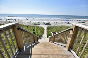 Eine Holztreppe führt zu einem Strand mit freiem Blick auf das Meer und Badegäste.