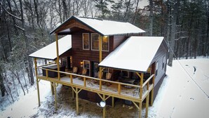 Une cabane en rondins dotée d’une terrasse et d’un porche abrité, entourée d’une forêt enneigée.