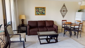 Una sala de estar con un sofá marrón, una mesa de centro de vidrio, un sillón estampado, una mesita con una lámpara, una mesa de comedor con sillas y un adorno en la pared.