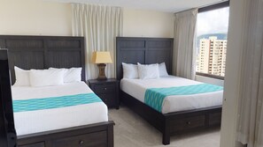 Habitación de hotel con dos camas, una mesita de noche con lámpara y vistas a edificios a través de la ventana.