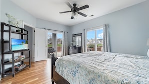 Un dormitorio con cama, ventilador de techo, televisor sobre un mueble y vista al exterior.