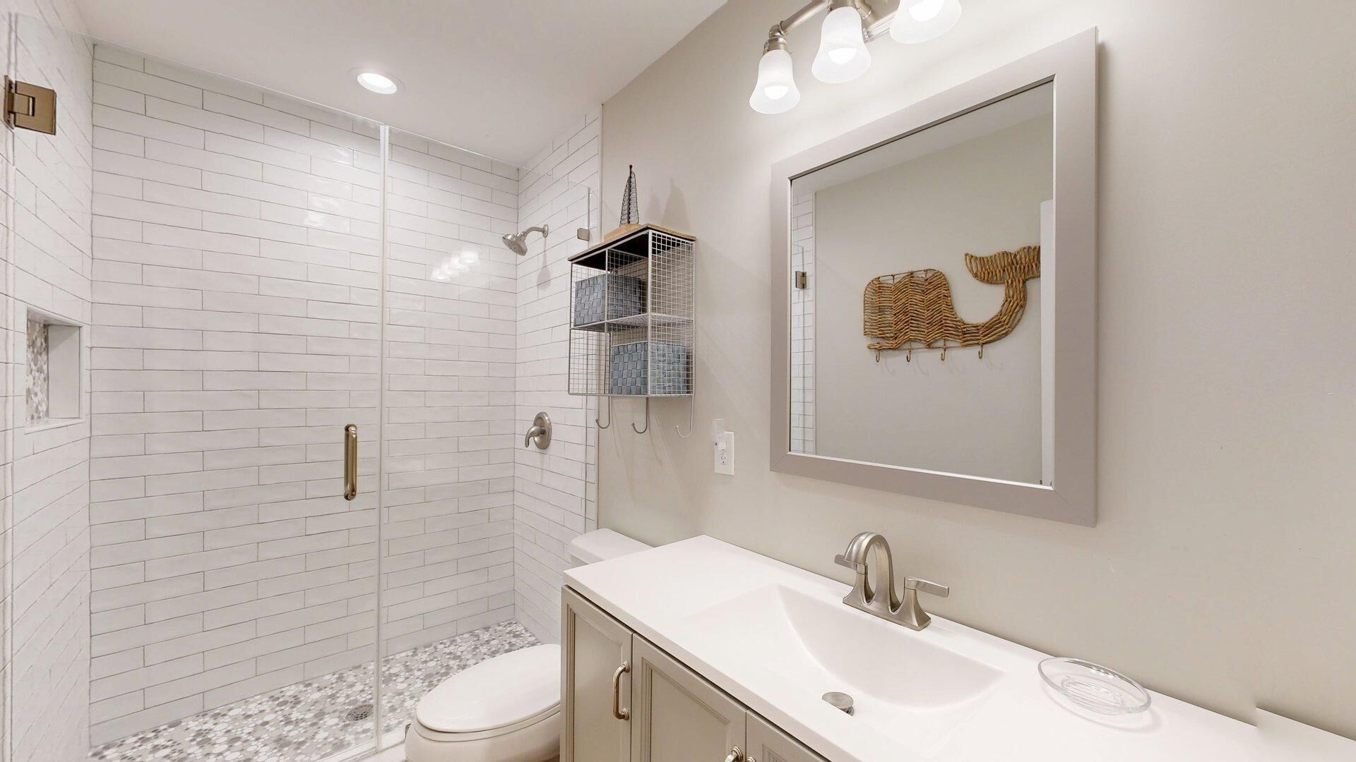 Une salle de bain moderne avec des carreaux de céramique blancs rectangulaires, une douche en verre, un lavabo blanc avec un miroir et des toilettes.