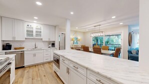 Une cuisine moderne avec des meubles blancs, un plan de travail en marbre et un coin repas doté de chaises en osier, offrant une vue sur la verdure.