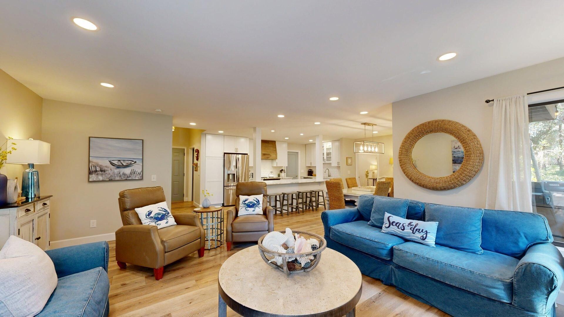 A spacious living room with a blue sofa, a brown leather chair, and a round coffee table. A kitchen and dining area are visible in the background.