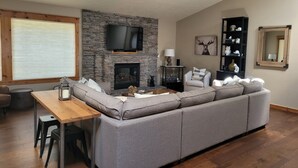 Een woonkamer met een stenen openhaard, een grote flatscreen-tv, een grijze modulaire bank, een houten salontafel, en een eethoek met een houten tafel en stoelen.
