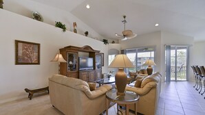 A spacious living room with a beige sofa, a glass coffee table, and a television set. There is a ceiling fan, a large window, and a balcony with chairs.