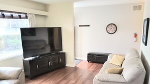 A living room with a flat-screen TV on a dark wooden cabinet, a grey sofa, and a matching ottoman. - Close to Everything A (Kelowna)