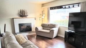 A living room with a fireplace, a sofa, a chair, and a television. - Close to Everything A (Kelowna)