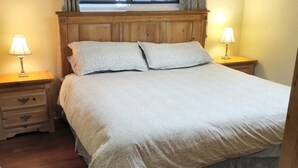 A bedroom with a wooden headboard, two bedside tables with lamps, a large bed with a grey duvet, and a window with curtains. - Close to Everything A (Kelowna)