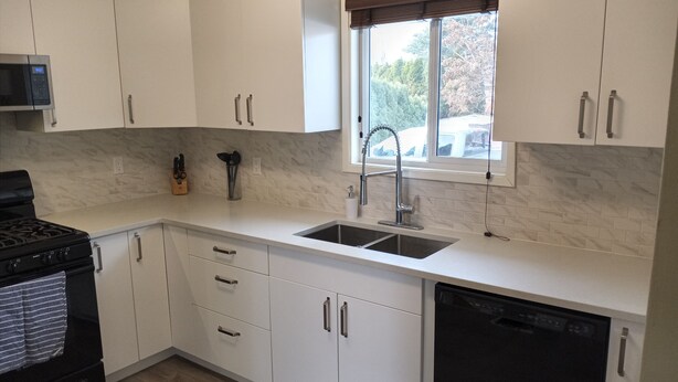 A modern kitchen with white cabinets, a black stove, a dishwasher, and a window with a view of trees. - Close to Everything A (Kelowna)
