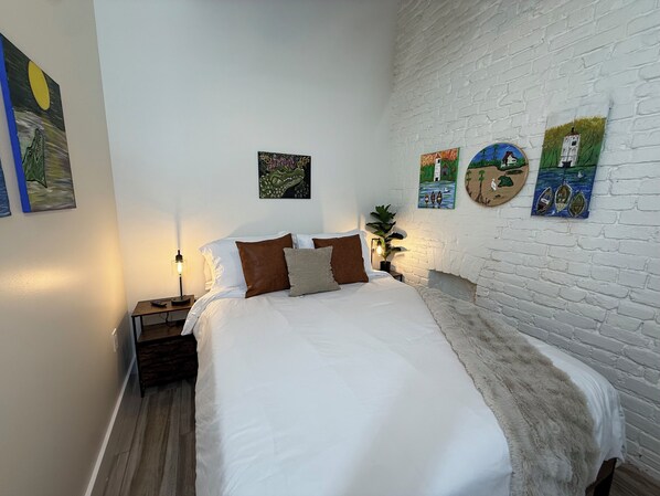 Un lit bien fait, avec une literie blanche et marron, un mur en briques blanches et plusieurs œuvres d’art accrochées au mur.