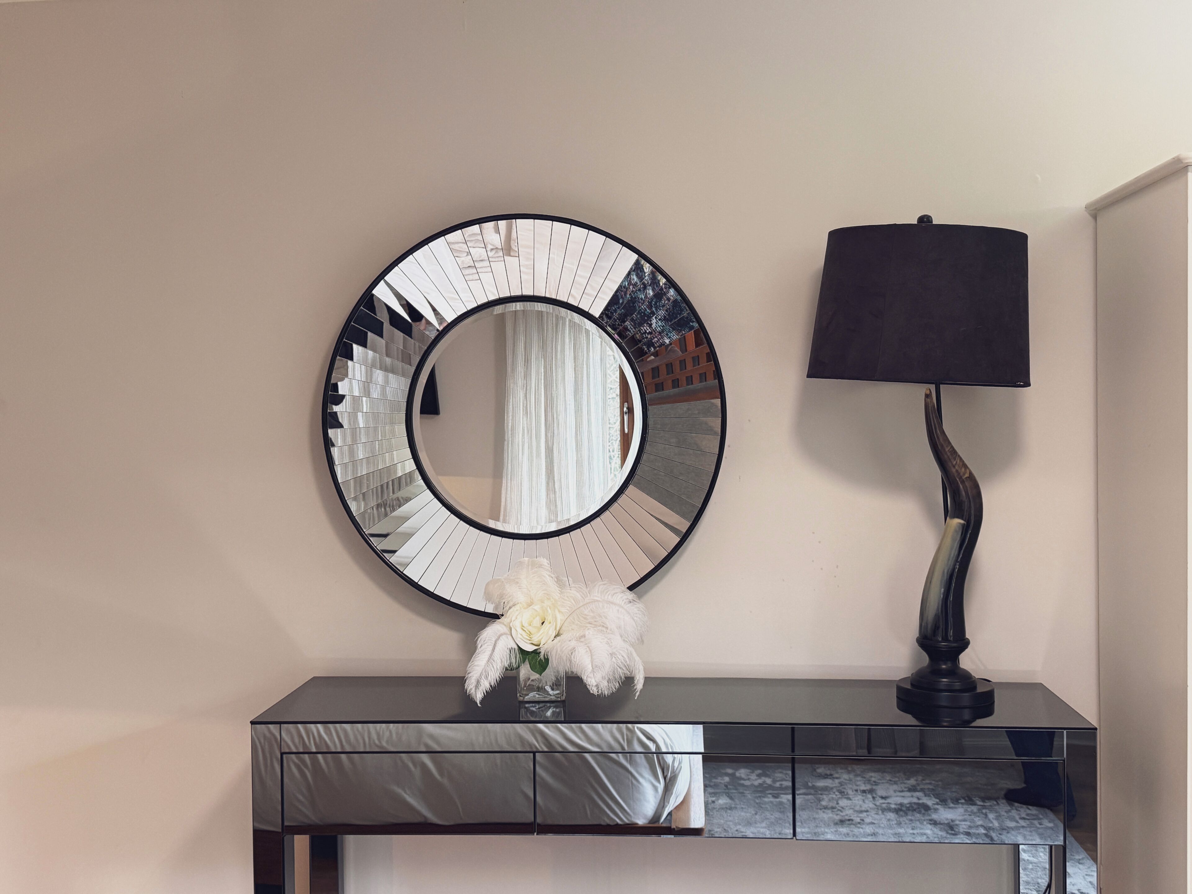Un miroir rond avec un cadre noir est accroché au mur, une lampe noire se trouve à côté, et un vase rempli de fleurs blanches est posé sur une console en dessous.